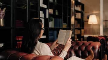 En person sitter i en lädersoffa och läser en bok i ett rum med en bokhylla och en lampa.