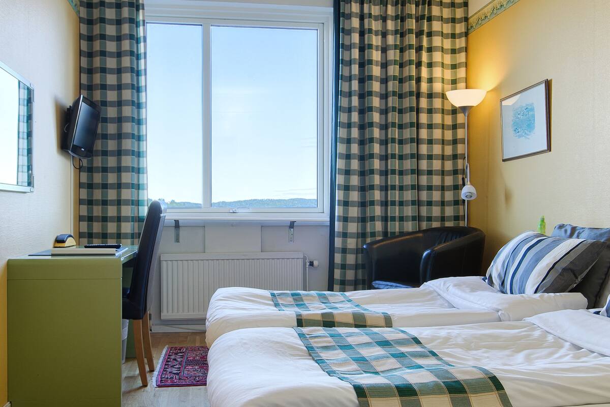 Une chambre d’hôtel avec un lit, un bureau, une chaise, une fenêtre avec des rideaux, une lampe et un tableau au mur.