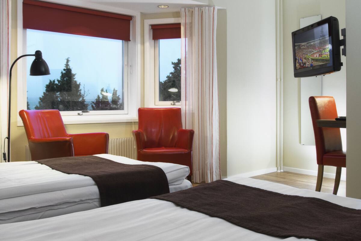 Une chambre d’hôtel avec deux lits, des chaises rouges, un téléviseur fixé au mur et une fenêtre donnant sur des arbres.