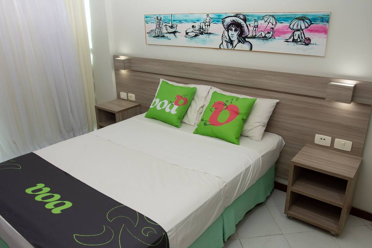 A neatly made bed with green and white bedding, a bedside table with a lamp, and a wall art depicting a beach scene.