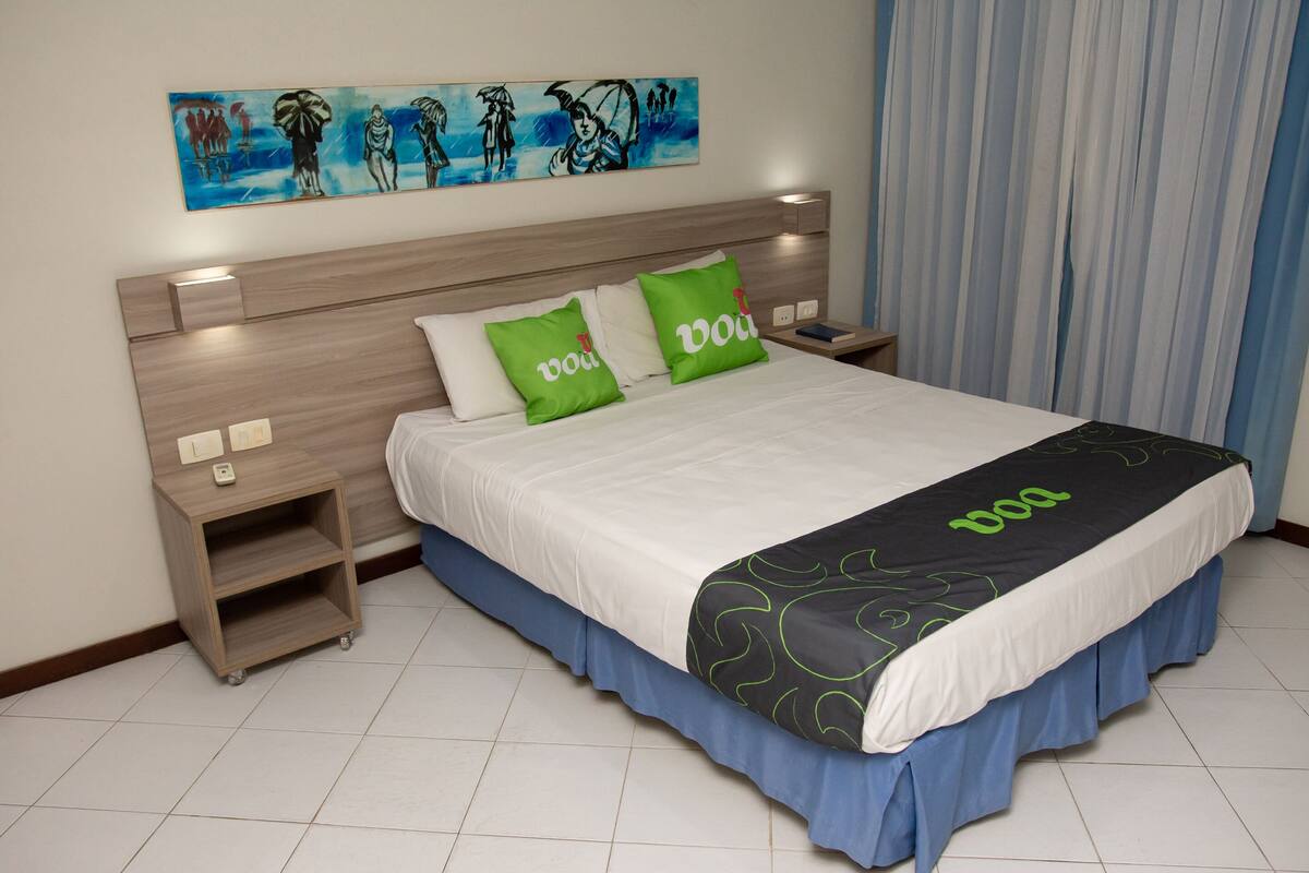 A neatly made bed with a white and blue bedspread, two green pillows, and a black and white blanket with 'VooDoo' branding.