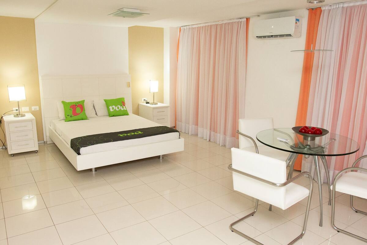 A hotel room with a bed, two bedside tables with lamps, a glass table with chairs, and a window with curtains.