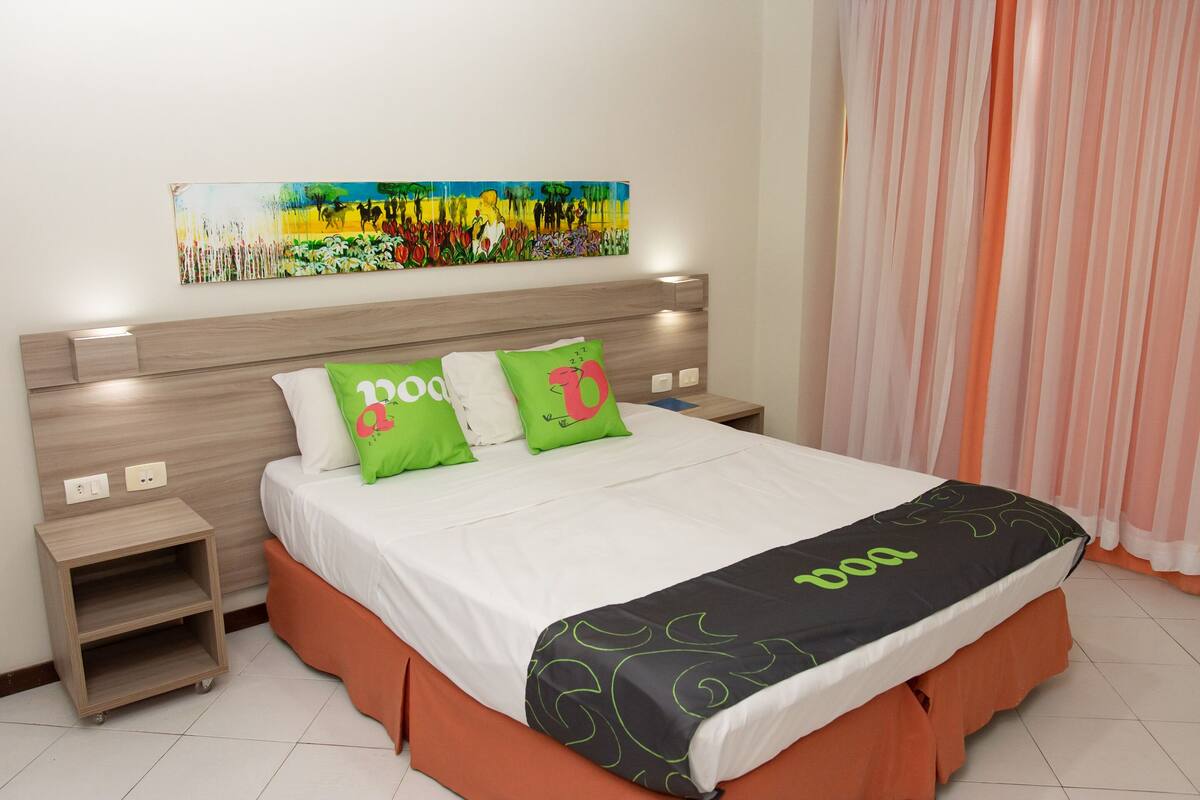 A neatly made bed with a white and orange bed skirt, two green pillows with a pink design, and a black and orange blanket with the word "doo" on it.