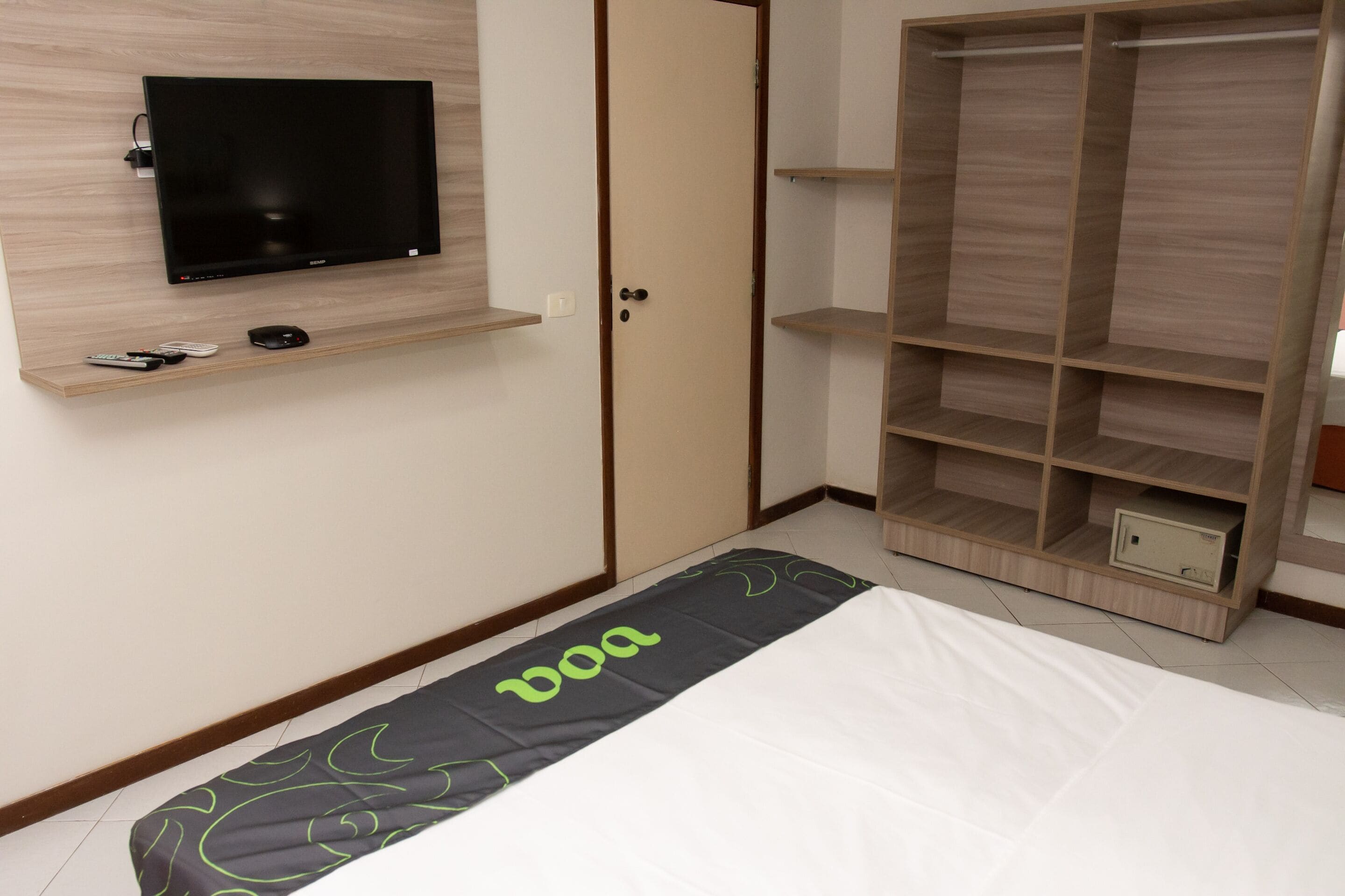 A bedroom with a bed, a TV mounted on the wall, a wardrobe, and a small shelf with remote controls.