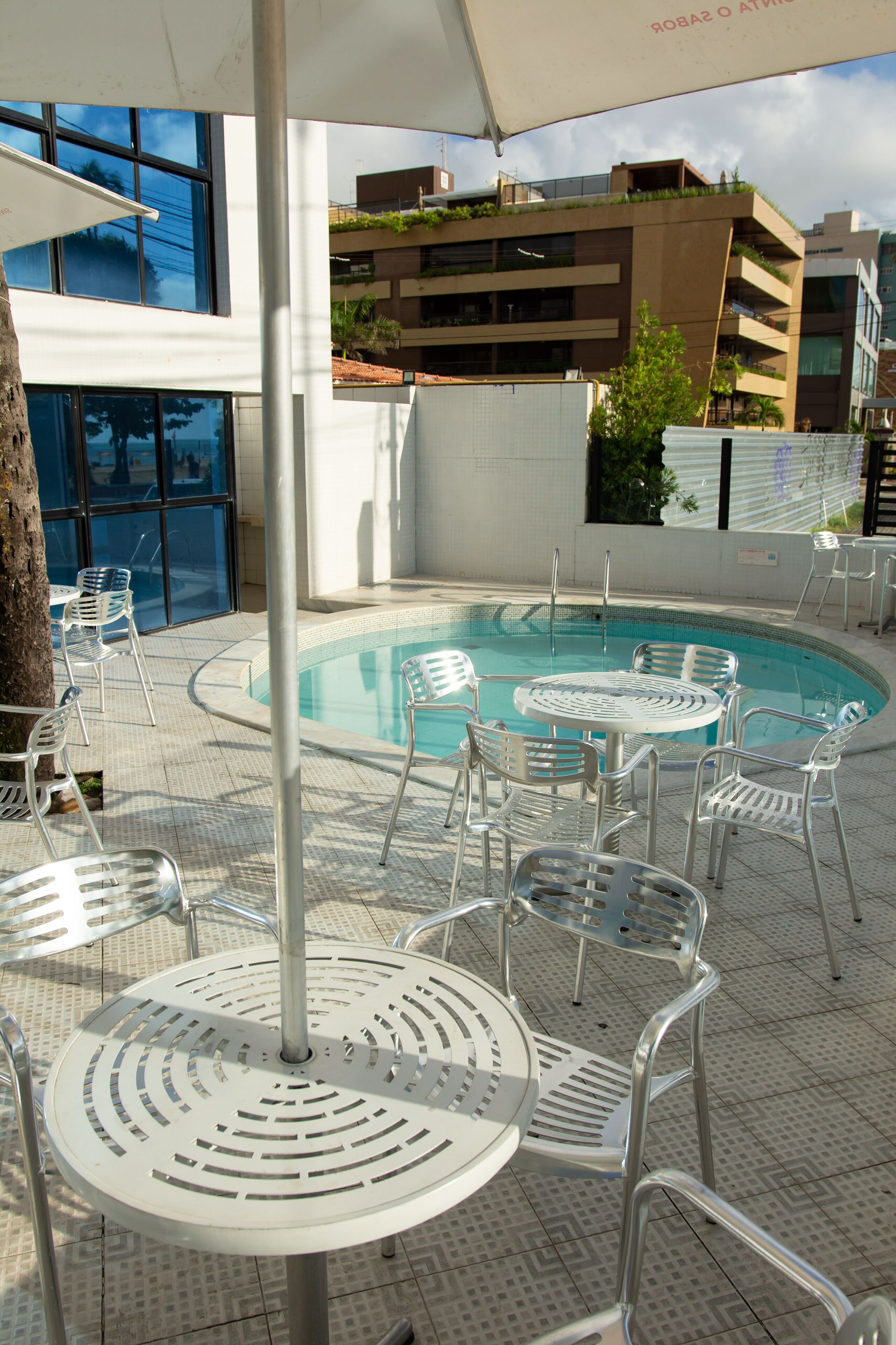An outdoor swimming pool area with metal chairs and tables, surrounded by buildings with balconies and large windows.