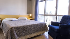 A hotel room with a large bed, a chair, a nightstand, and a window with a view of buildings. - Like U Hotel Brasilia (Brasília)