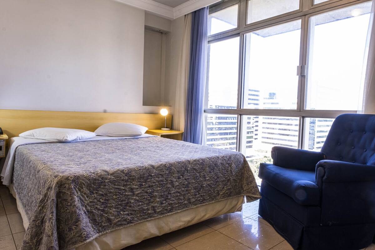 Quarto de hotel com uma cama grande, uma cadeira, uma mesinha de cabeceira e uma janela com vista para edifícios.