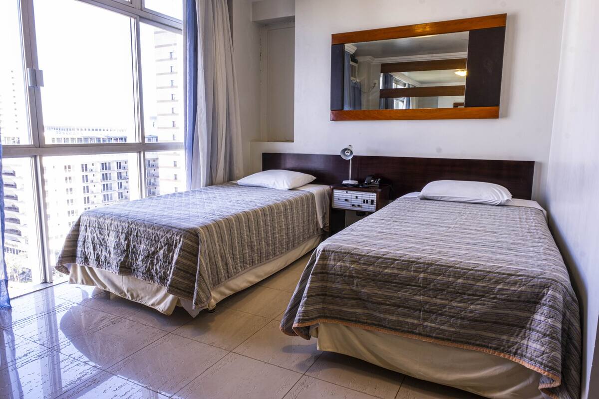 Quarto de hotel com duas camas, uma janela grande e um espelho na parede.