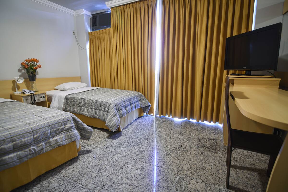 Quarto de hotel com duas camas, uma mesa, uma televisão e uma janela com cortinas.