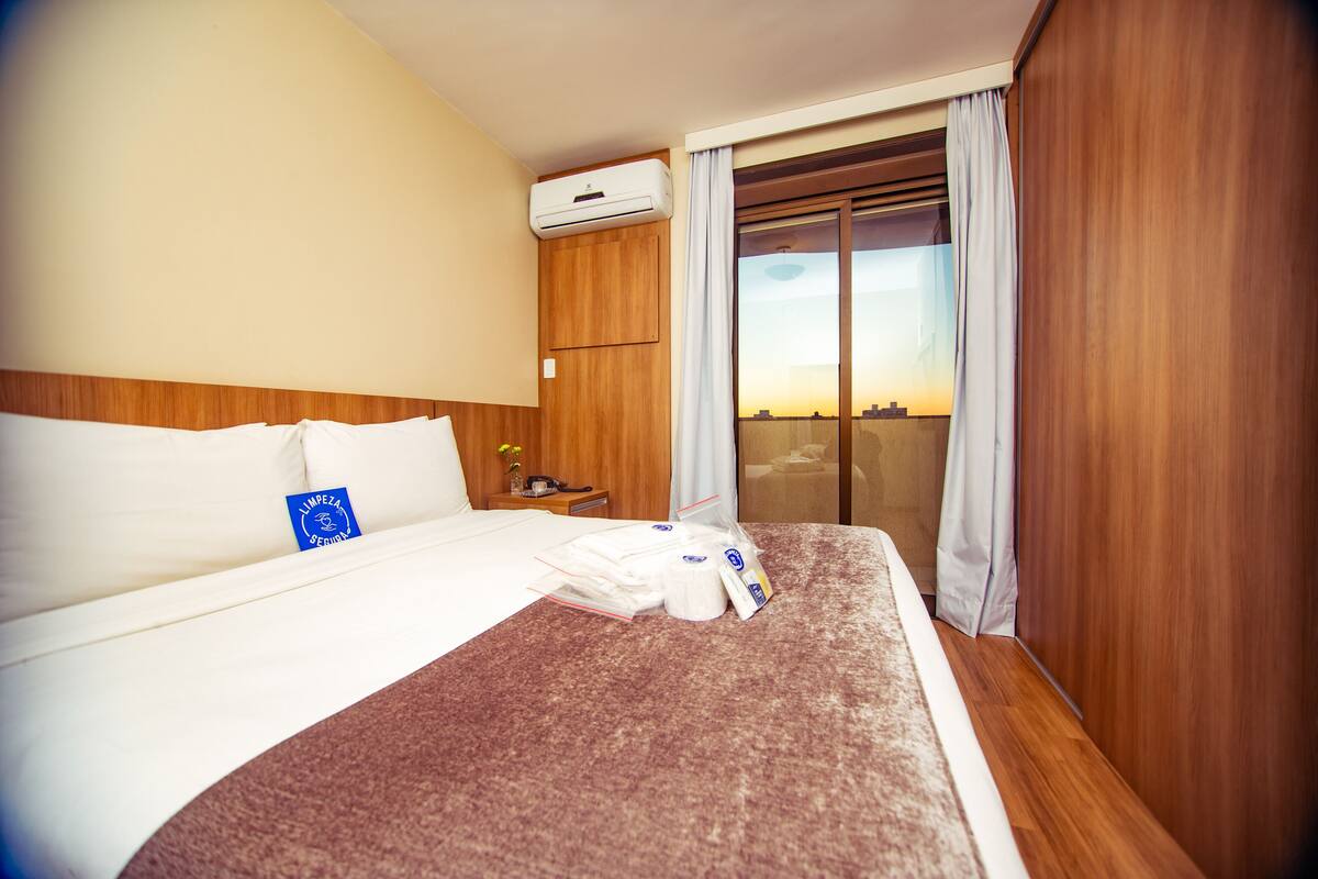 Ein Hotelzimmer mit Bett, Nachttisch, Klimaanlage, einem Fenster mit Vorhängen und Blick auf die Stadt.