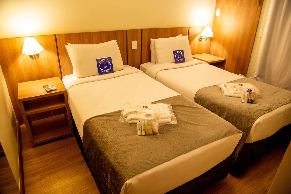 Habitación de hotel con dos camas, cada una con cabecero de madera, ropa blanca y una mesita con lámpara. Hay toallas y artículos de aseo en las camas.