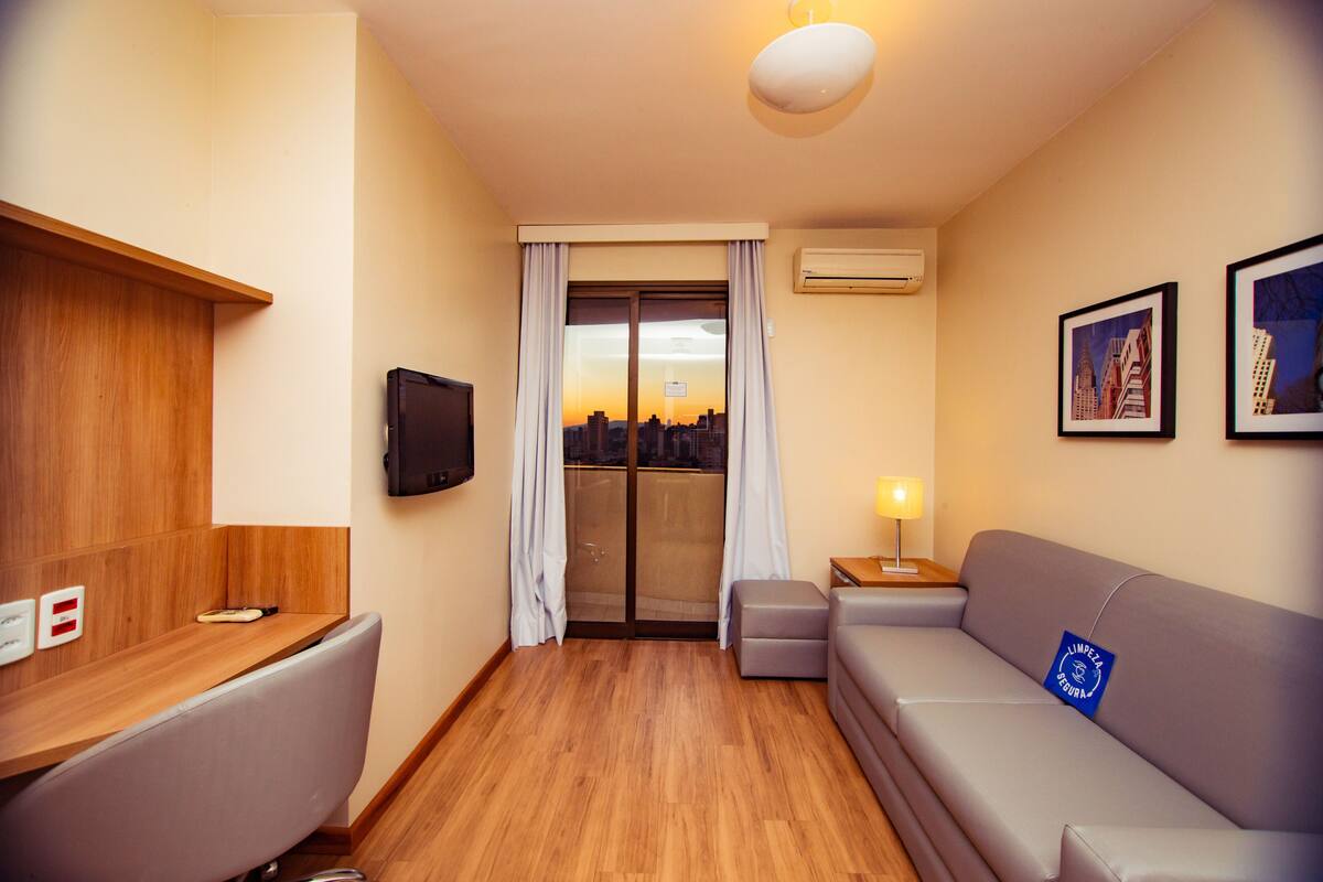 Ein Hotelzimmer mit einem Holztisch, einem grauen Sofa, einem kleinen Tisch mit Lampe, einem Fernseher und einer Glastür mit Vorhängen.