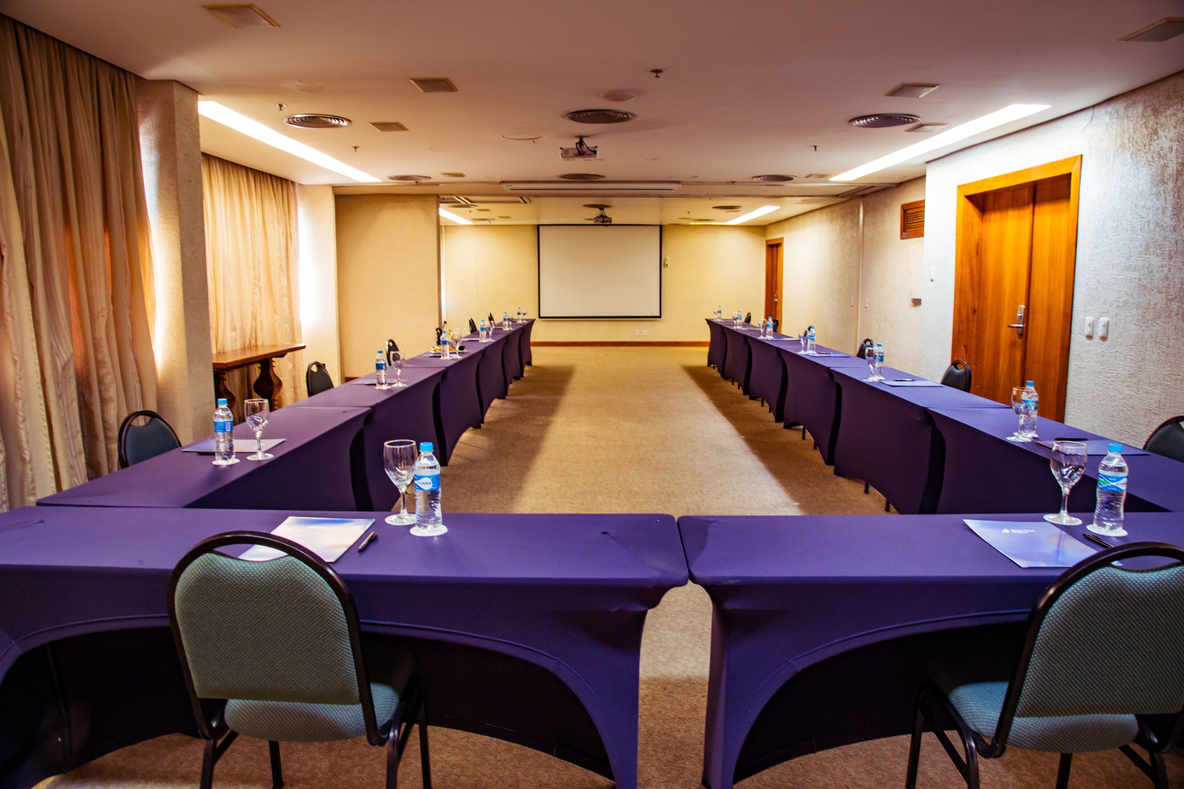 Un salón de conferencias con una larga mesa, sillas, botellas de agua y una pantalla de proyector.