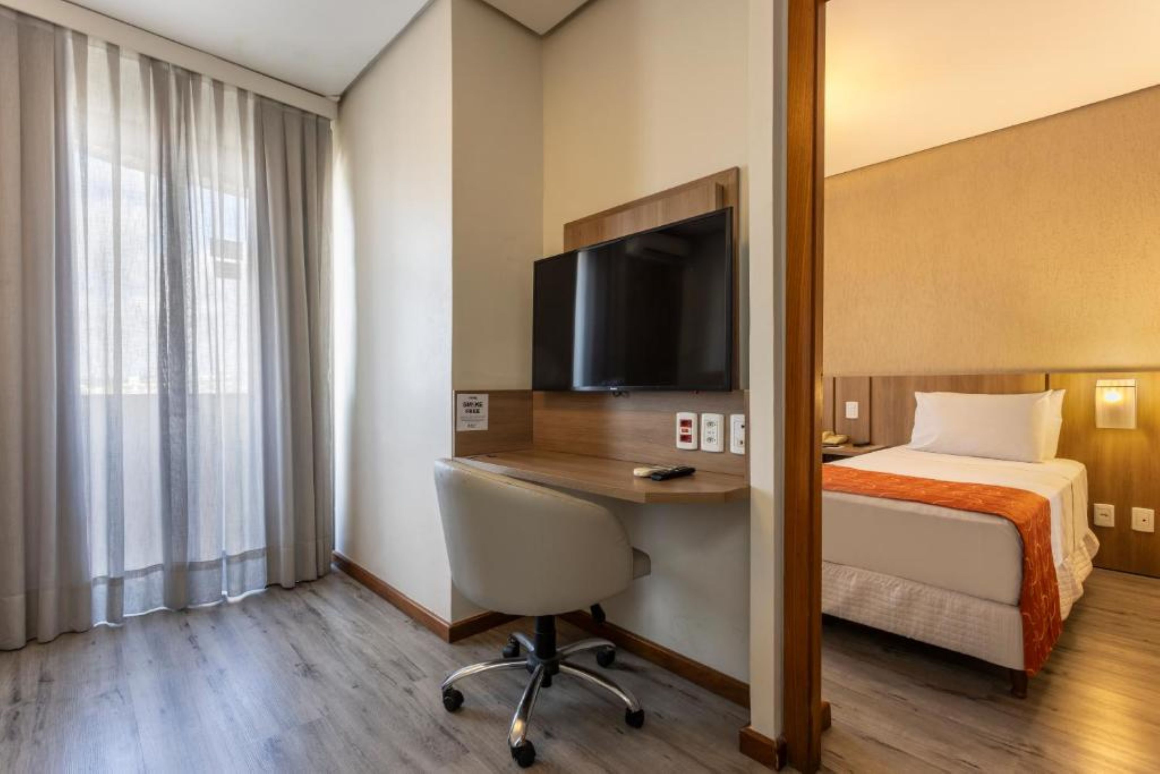 A hotel room with a bed, a desk with a chair, a television, and a window with curtains.
