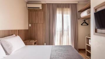Habitación de hotel con cama, mesita de noche, aire acondicionado, ventana con cortinas y televisor montado en la pared.