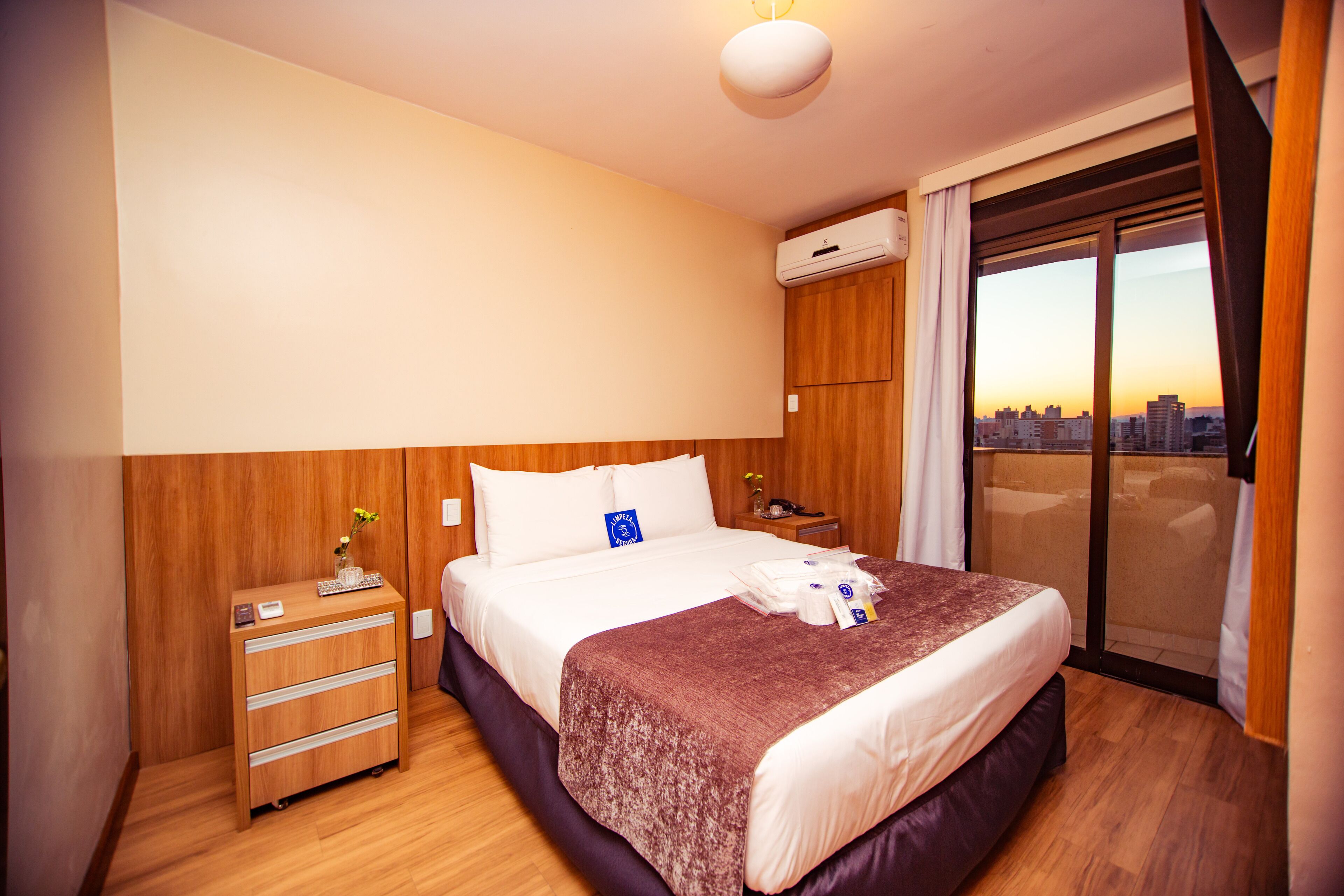 Quarto de hotel com uma cama grande, uma cômoda de madeira, uma mesa de cabeceira e vista da cidade através de uma porta de vidro deslizante.