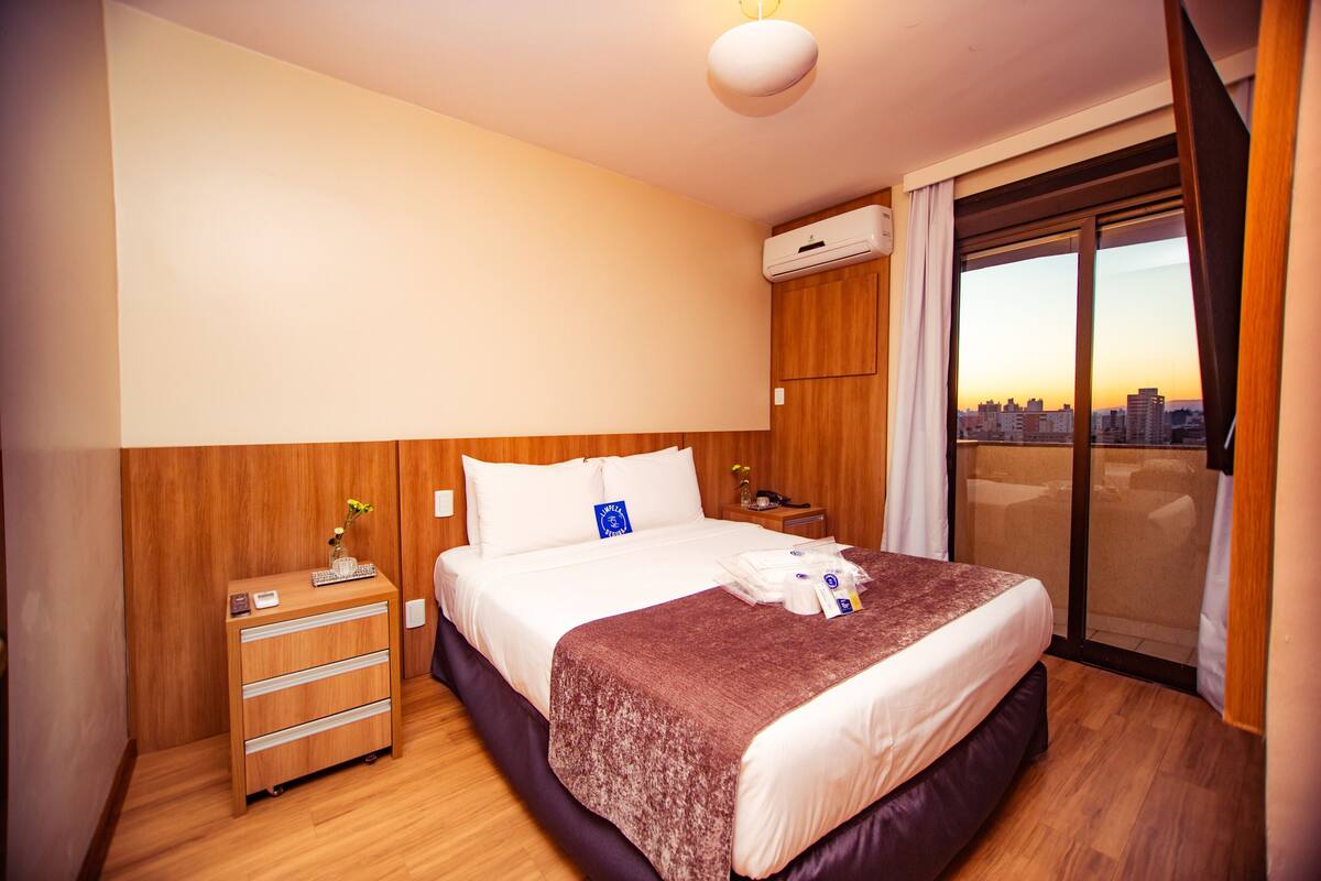 Habitación de hotel con una cama grande, un aparador de madera, una mesita de noche y vistas de la ciudad a través de una puerta corrediza de vidrio.