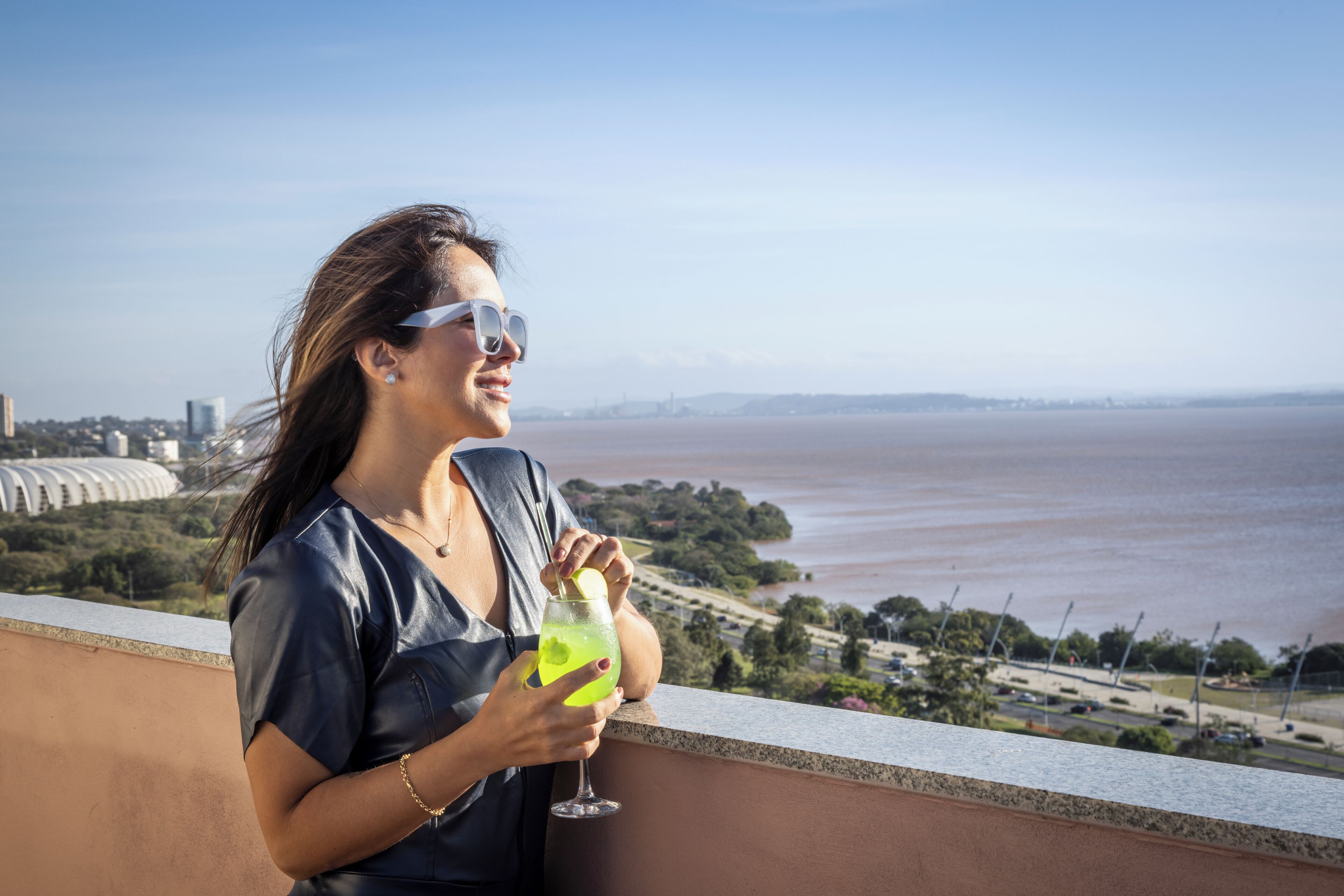 Una persona sosteniendo una bebida en un balcón con vistas a un río y al paisaje urbano.