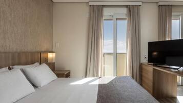 A hotel room with a large bed, a TV on a wooden cabinet, and a view of the sky through the window.