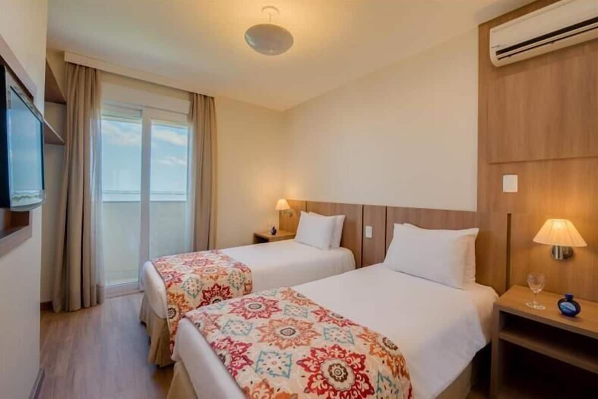 Two single beds with white duvets and colorful throw blankets, a flat-screen TV, and a nightstand with a lamp.