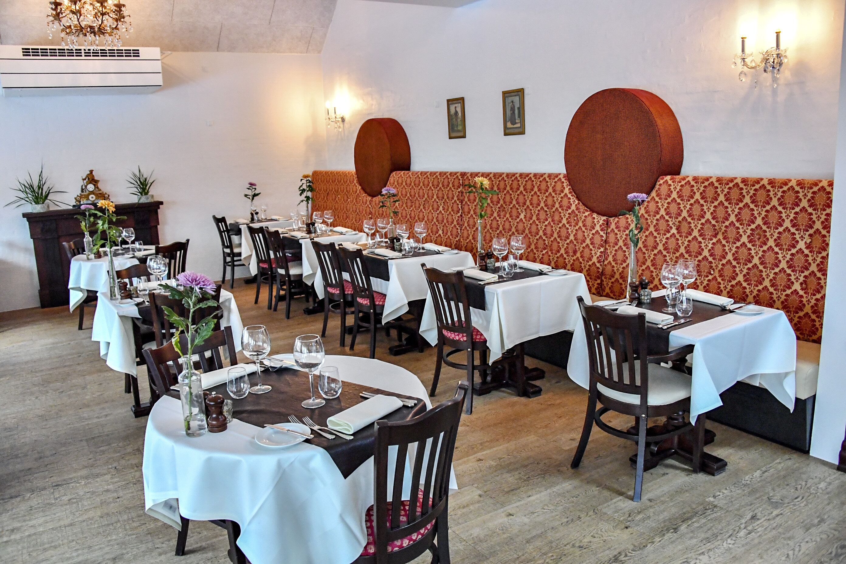 A restaurant with round tables, white tablecloths, and wooden chairs.