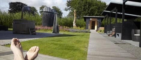 A wooden deck leading to a building with a covered outdoor area, a small structure, and a tall chimney.
