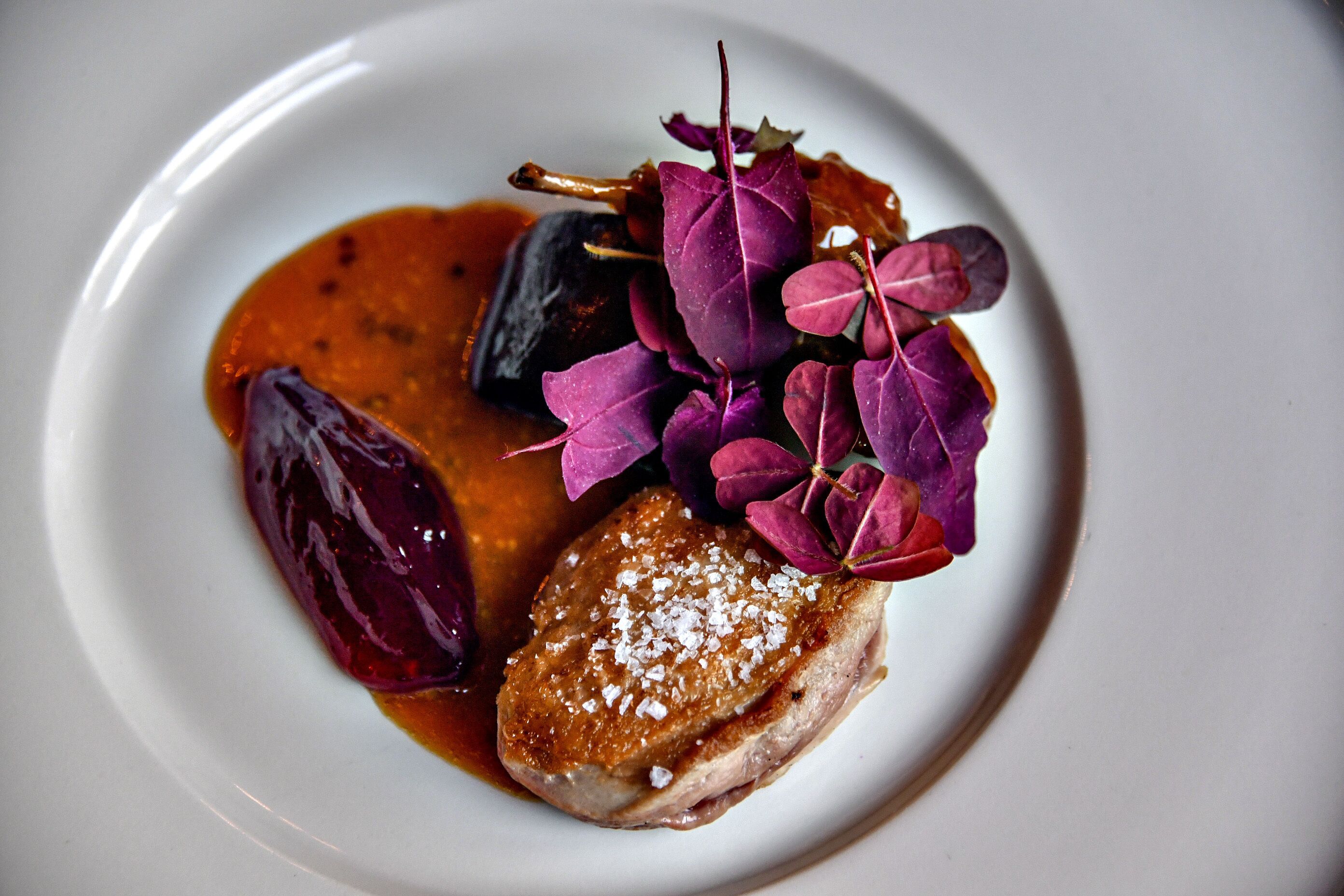 Une assiette contenant un morceau de viande, un légume foncé et des feuilles violettes.