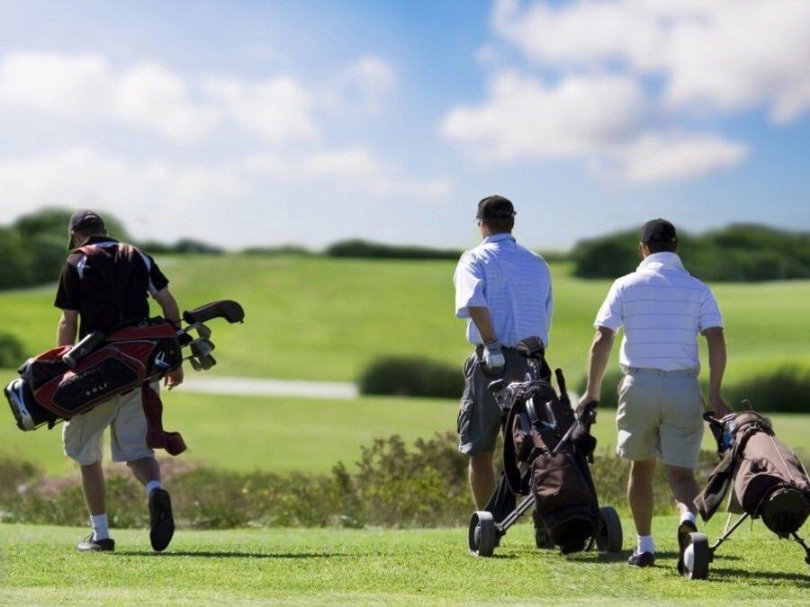 Tres personas caminando en un campo de golf, cargando bolsas de golf y acompañadas por un carrito de golf.