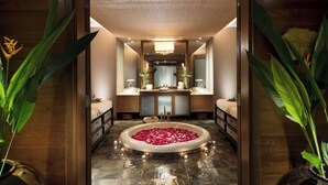 Un baño lujoso con una bañera central rodeada de pétalos de rosa, un tocador de madera con espejo y dos lavamanos.