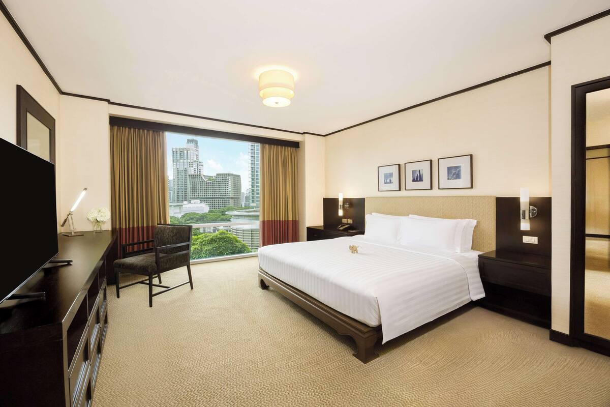 Habitación de hotel con una cama grande, un escritorio con televisor, una silla y vista a la ciudad por la ventana.