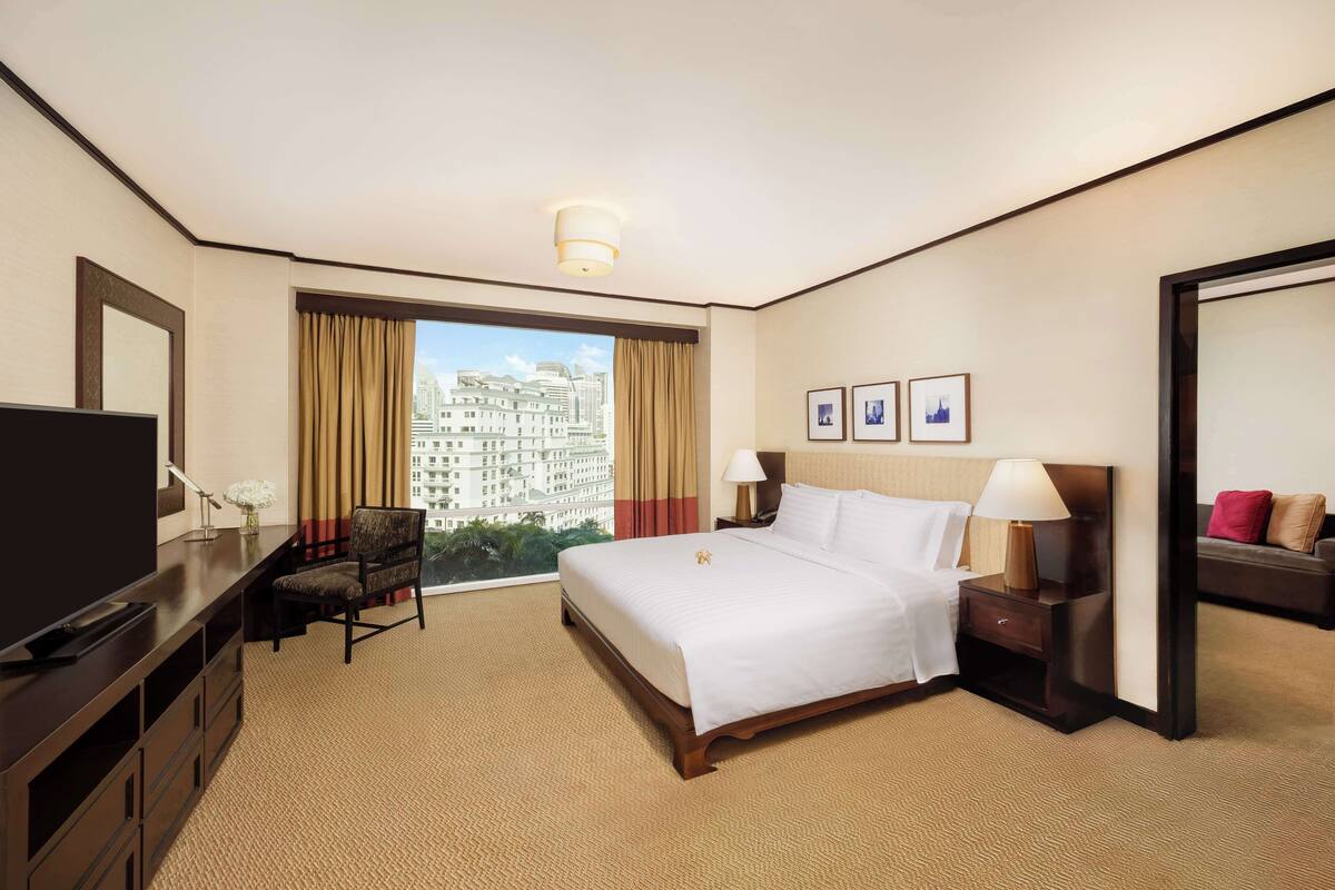 Habitación de hotel con una cama grande, un escritorio con televisor, una silla, una mesita de noche con lámparas y vistas a un paisaje urbano por la ventana.