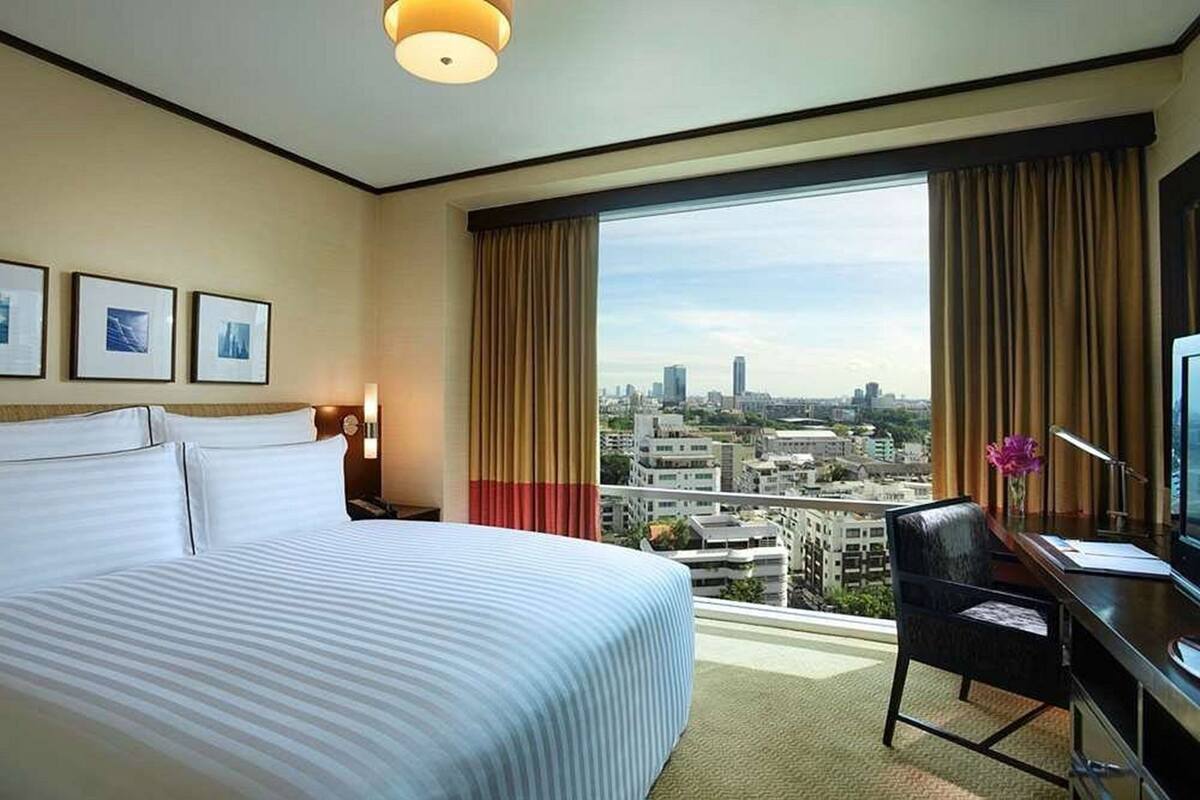 Habitación de hotel con una cama grande, un escritorio con computadora, una silla y vistas a un paisaje urbano a través de un ventanal amplio.
