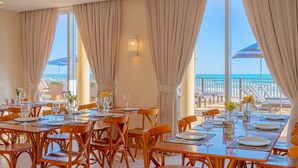 Un comedor con mesas dispuestas para huéspedes, ventanas grandes con cortinas y vista a la playa.