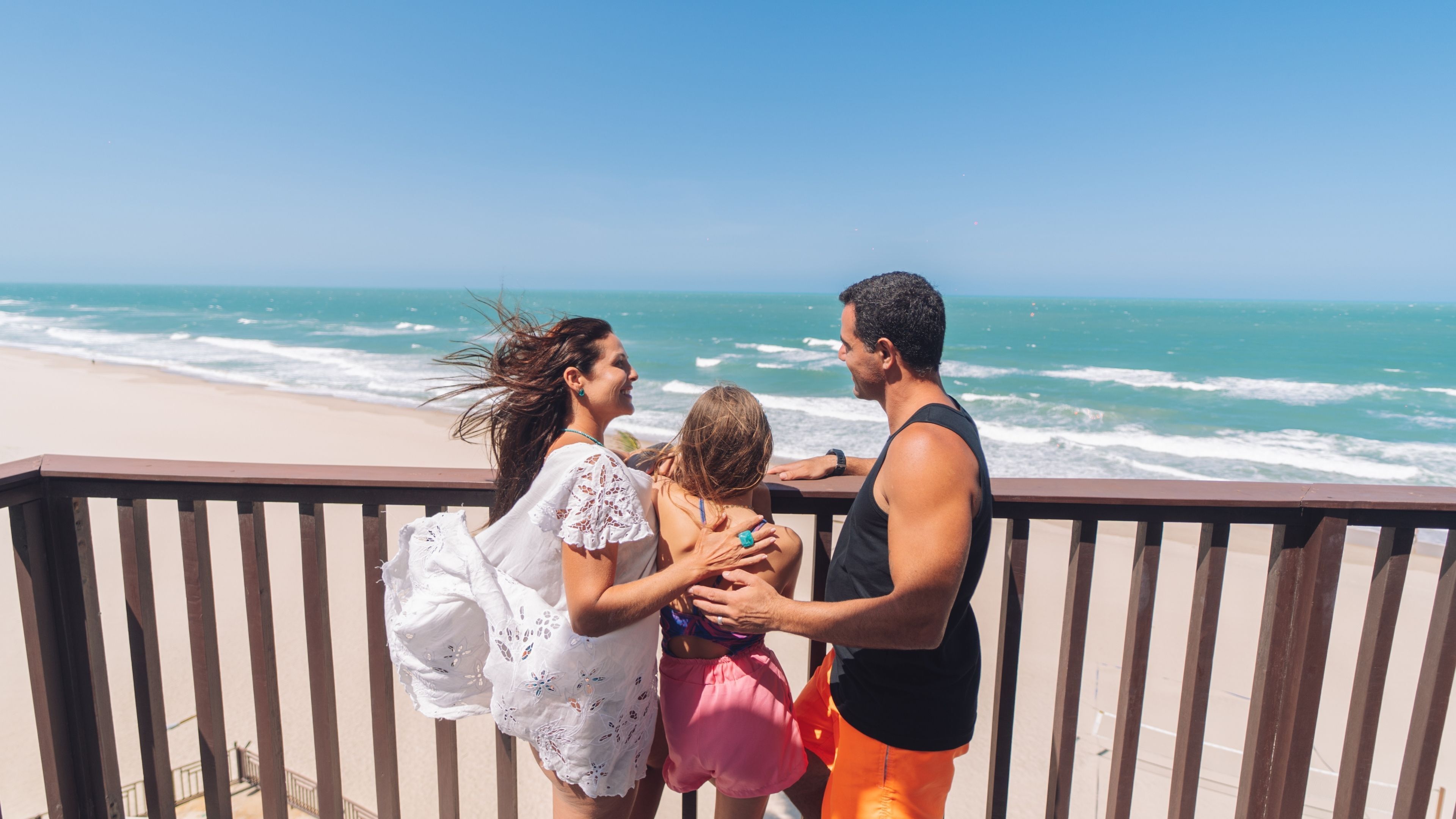 Varanda com vista para uma praia, ondas, guarda-corpo e céu claro.