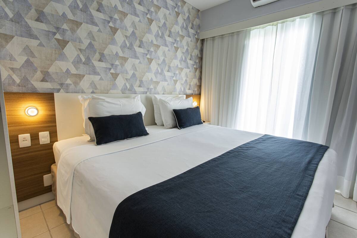Hotellihuone, jossa on sänky, kaksi yöpöytää, geometrisesti kuvioitu seinä ja ikkuna, jossa on verhot.