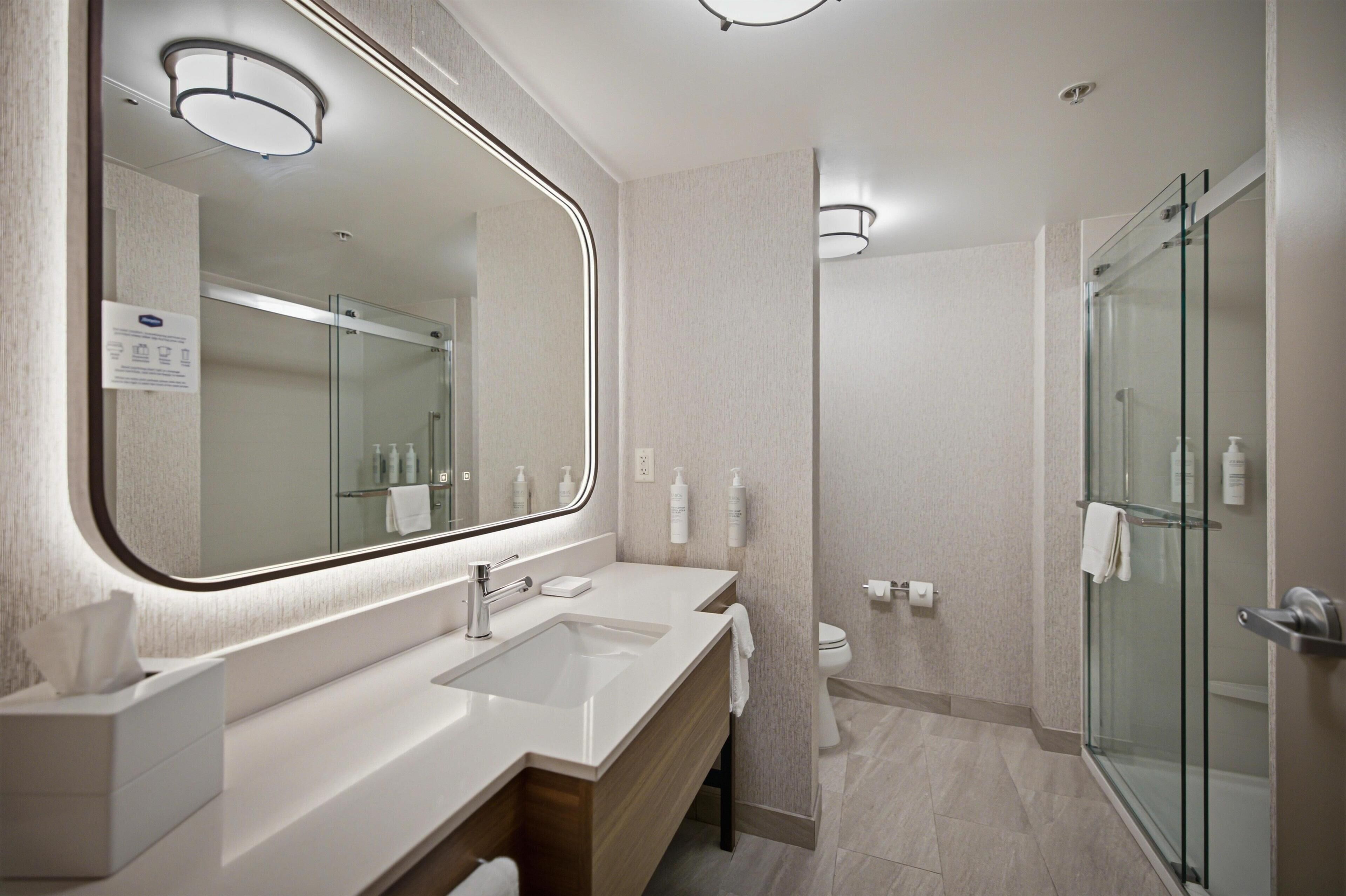 A modern bathroom with a large mirror, dual sinks, a glass shower enclosure, and a toilet.