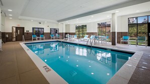 A swimming pool with clear blue water, surrounded by lounge chairs and tables, and a view of greenery outside.