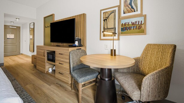 A hotel room with a bed, a desk with a flat-screen TV, a microwave, and a coffee maker. There are two upholstered chairs and a small round table. The room has wooden flooring and a large mirror on the wall.