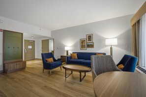 Habitación de hotel con un sofá azul, dos sillones, una mesita de centro redonda y una lámpara. Se observan cuadros en la pared y una puerta con espejo.
