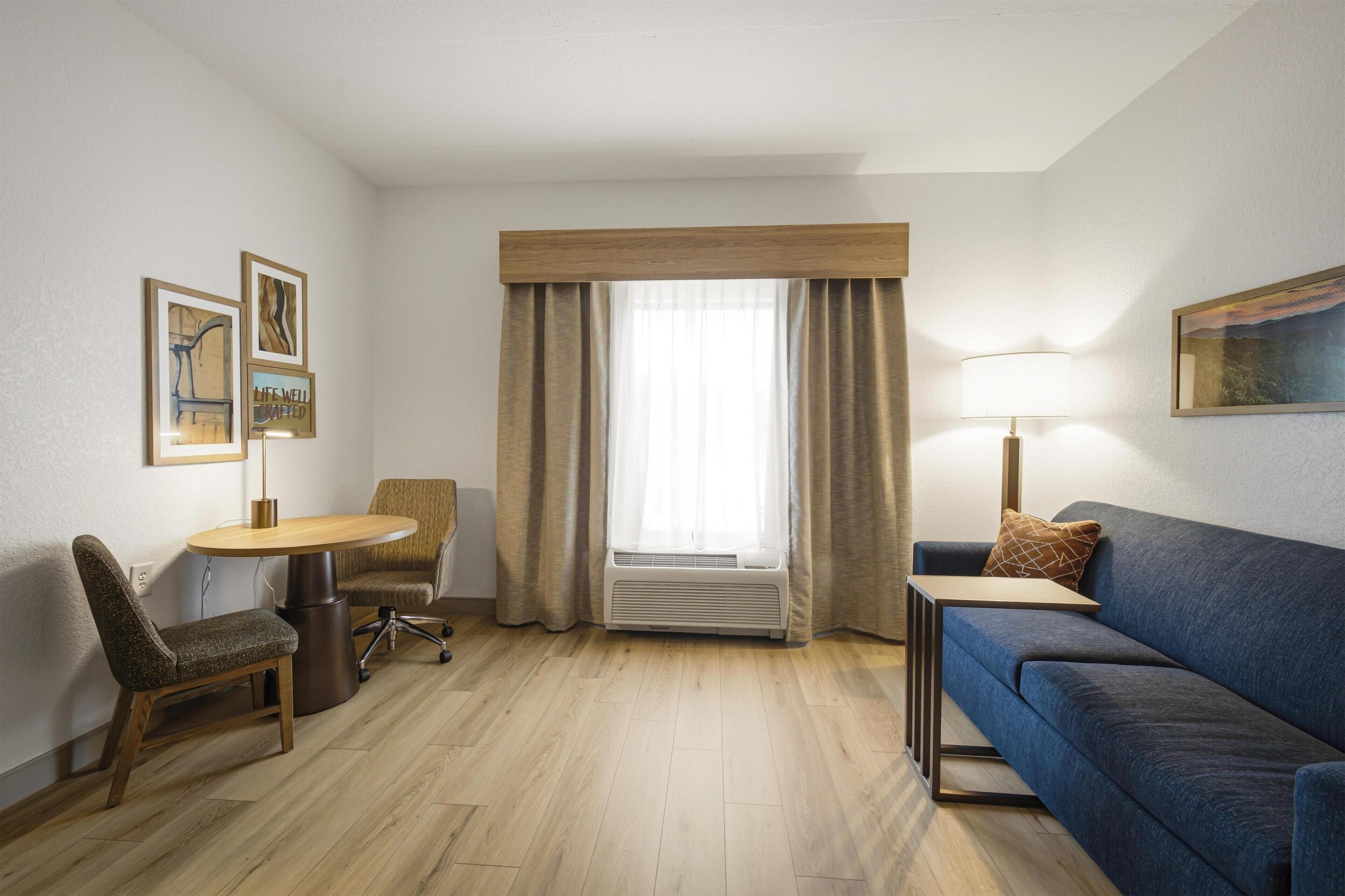 Habitación de hotel con piso de madera, sofá azul, una mesita, una silla, escritorio, lámpara y ventana con cortinas.