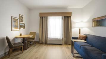 Habitación de hotel con piso de madera, sofá azul, una mesita, una silla, escritorio, lámpara y ventana con cortinas.