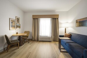 Habitación de hotel con piso de madera, sofá azul, una mesita, una silla, escritorio, lámpara y ventana con cortinas.