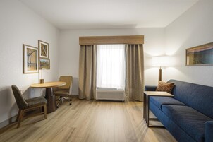 Habitación de hotel con piso de madera, sofá azul, una mesita, una silla, escritorio, lámpara y ventana con cortinas.