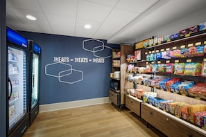 Interior de un supermercado con un congelador a la izquierda, secciones de aperitivos y alimentos congelados, y una pared con patrones hexagonales y la palabra 'TREAT' repetida.
