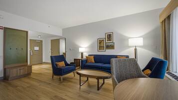Habitación de hotel con un sofá azul, dos sillones, una mesita de centro redonda y una lámpara. Se observan cuadros en la pared y una puerta con espejo.