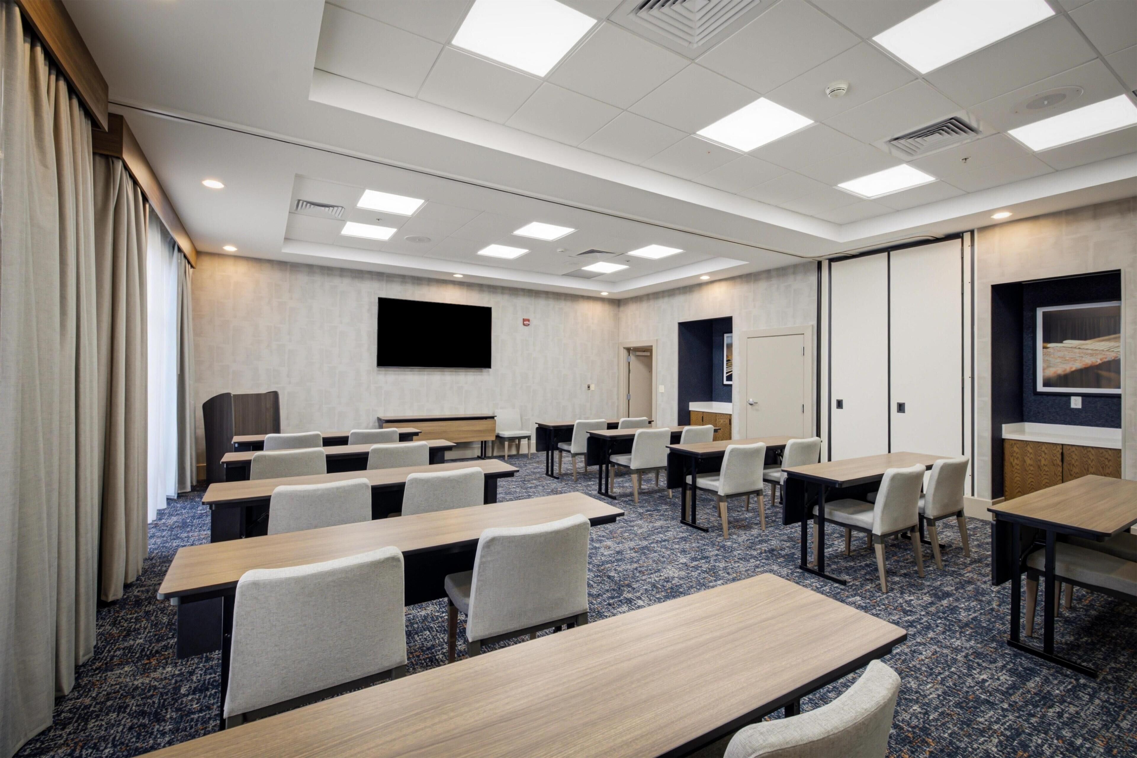 A conference room with multiple tables and chairs, a flat-screen TV mounted on the wall, and a door leading to another room.