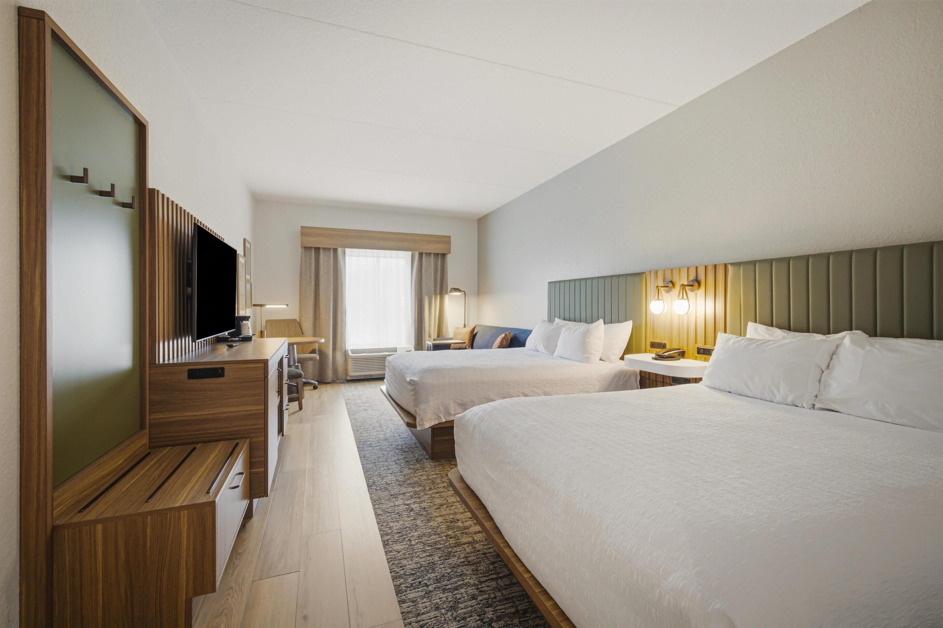 A hotel room with two beds, a TV, a desk, and a window with curtains.