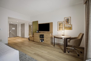 A hotel room with a bed, a desk with a computer, a chair, a TV, and artwork on the wall.
