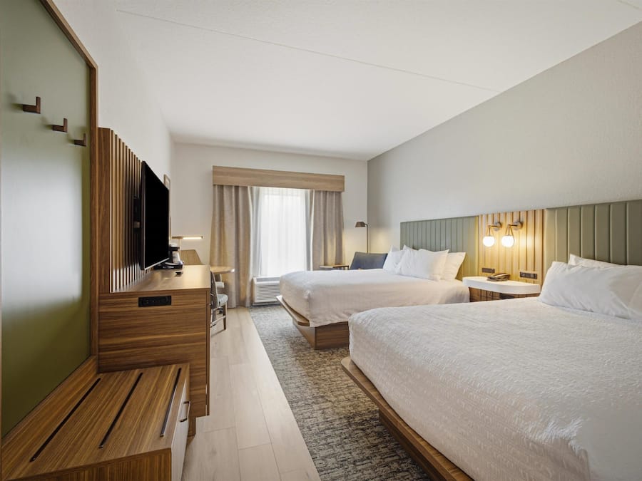 A hotel room with two beds, a desk, a television, and a window with curtains.