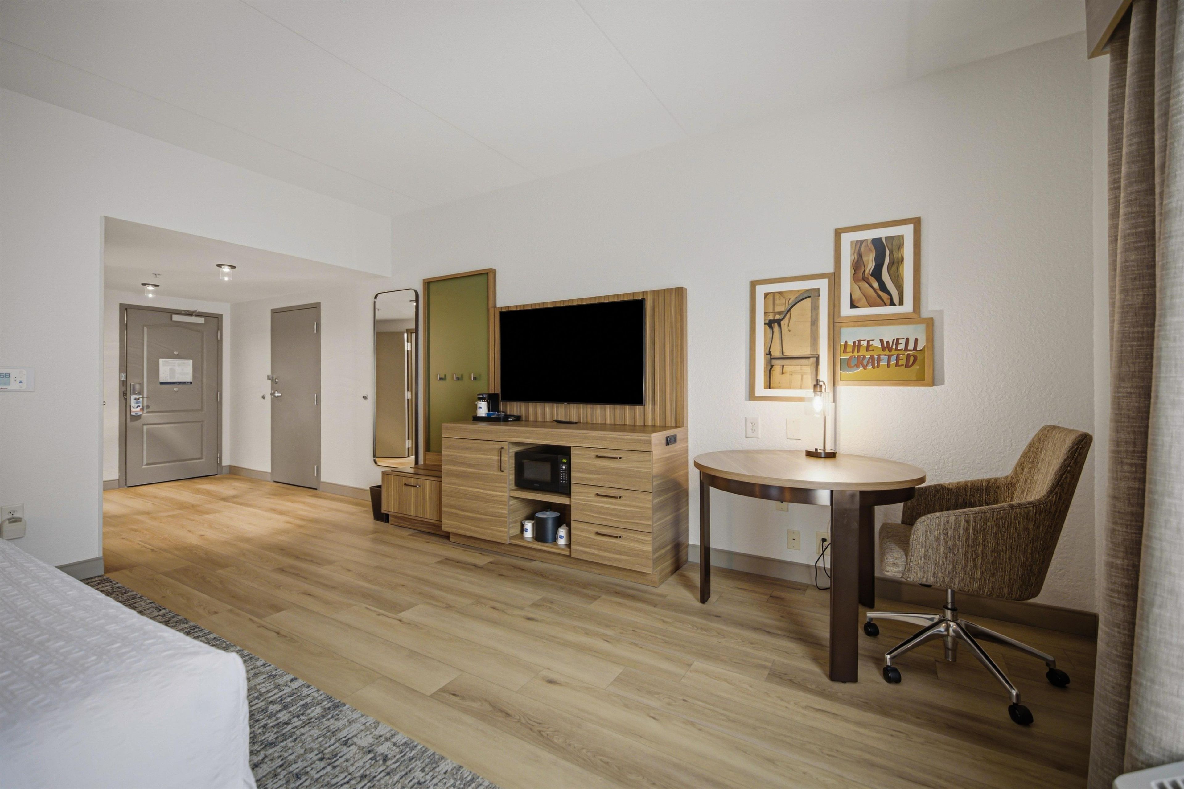 A hotel room with a bed, a desk with a computer, a chair, a TV, and artwork on the wall.